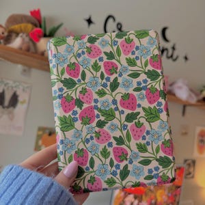 May include: A rectangular case with a cream background, covered in a pattern of pink strawberries, blue flowers, and green leaves. The case is held in a person's hand, and the background is a blurred room with a shelf and wall decor.