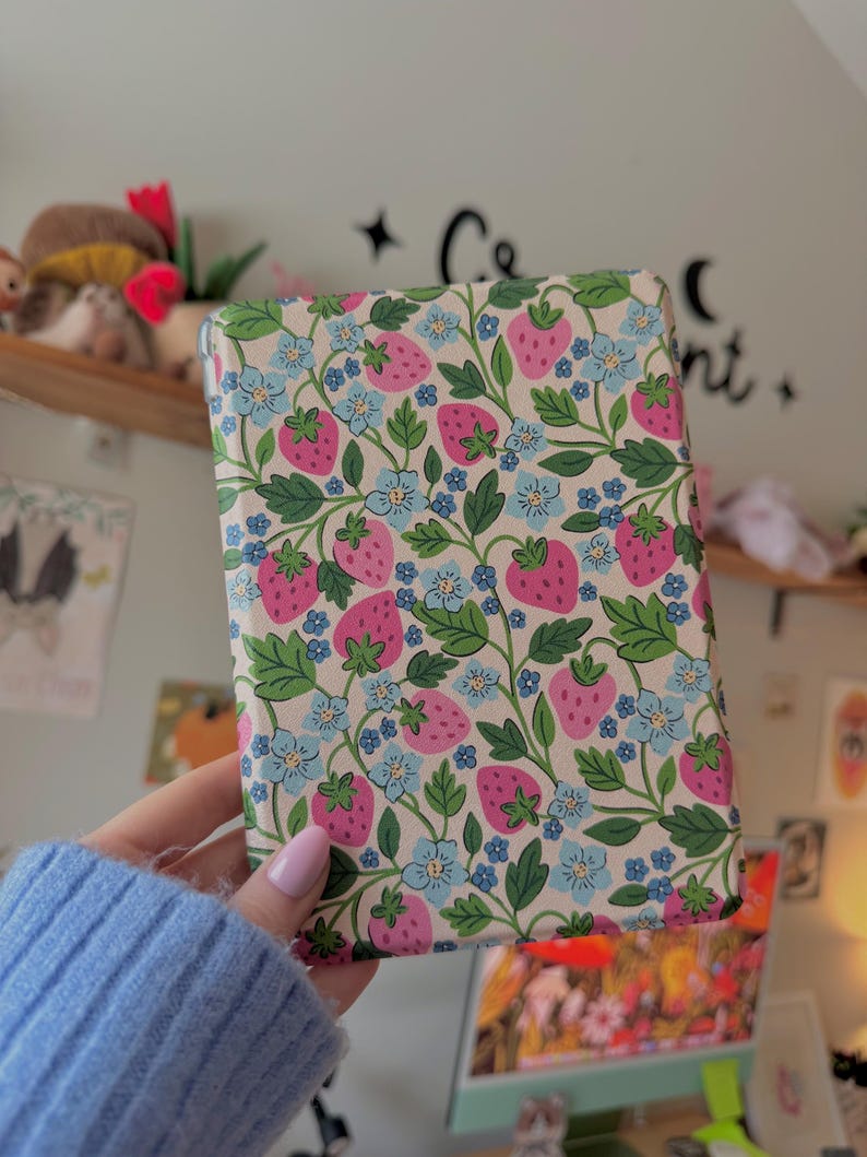 May include: A rectangular case with a cream background, covered in a pattern of pink strawberries, blue flowers, and green leaves. The case is held in a person's hand, and the background is a blurred room with a shelf and wall decor.
