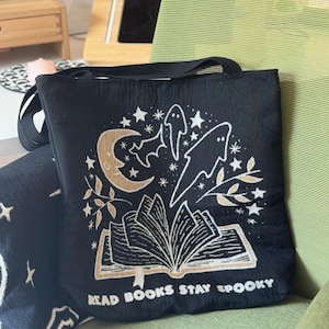 Puede incluir: Bolso tote negro con un dise&ntilde;o espeluznante que presenta una luna creciente, estrellas, murci&eacute;lagos y fantasmas. El dise&ntilde;o incluye un libro abierto con el texto "READ BOOKS STAY SPOOKY" en color dorado. El bolso tiene una correa negra.