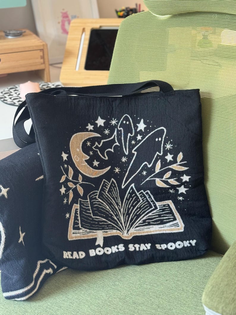 Puede incluir: Bolso tote negro con un dise&ntilde;o espeluznante que presenta una luna creciente, estrellas, murci&eacute;lagos y fantasmas. El dise&ntilde;o incluye un libro abierto con el texto "READ BOOKS STAY SPOOKY" en color dorado. El bolso tiene una correa negra.