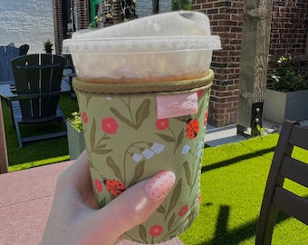 May include: A green fabric coffee cup sleeve with a floral and ladybug pattern. The sleeve is wrapped around a clear plastic cup with a lid. The background includes outdoor seating and a brick building.