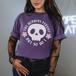May include: A purple t-shirt with rolled sleeves displaying a white skull graphic and the text "THE HORRORS PERSIST BUT SO DO I." Two small flowers are on either side of the skull. This is a clothing item.