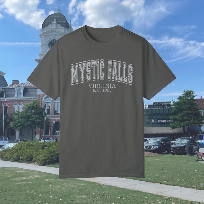 Puede incluir: Una camiseta gris oscura con un estampado gr&aacute;fico blanco que dice "Mystic Falls, Virginia Est. 1864".