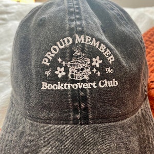 May include: A black denim baseball cap with white embroidery that reads "Proud Member of the Booktrovert Club". The embroidery also includes a small illustration of a stack of books with stars.