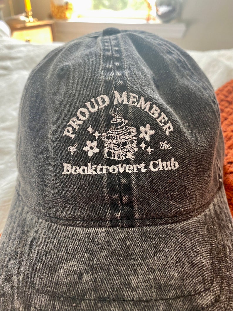 May include: A black denim baseball cap with white embroidery that reads "Proud Member of the Booktrovert Club". The embroidery also includes a small illustration of a stack of books with stars.
