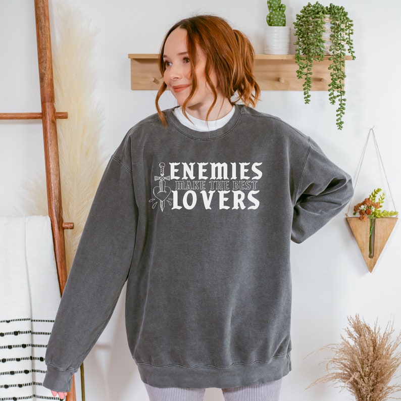 May include: A charcoal grey sweatshirt featuring the white text "ENEMIES MAKE THE BEST LOVERS". The design includes a sword, heart, and dagger. The sweatshirt is a relaxed fit.