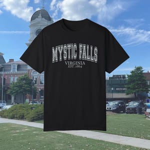 Puede incluir: Camiseta negra con un estampado gr&aacute;fico blanco que dice "Mystic Falls, Virginia Est. 1864".