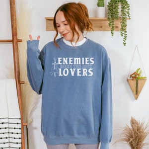 May include: A blue sweatshirt featuring the white text "ENEMIES MAKE THE BEST LOVERS". The design includes a sword and heart graphic. The sweatshirt is a relaxed fit.