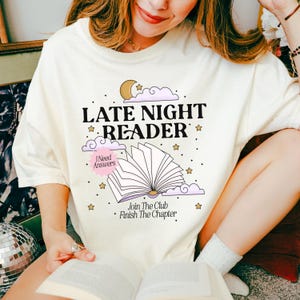 May include: Cream-colored t-shirt with the text "LATE NIGHT READER" in black, with a graphic of an open book, clouds, stars, and a moon. The shirt also has the phrases "I Need Answers" and "Join The Club Finish The Chapter".