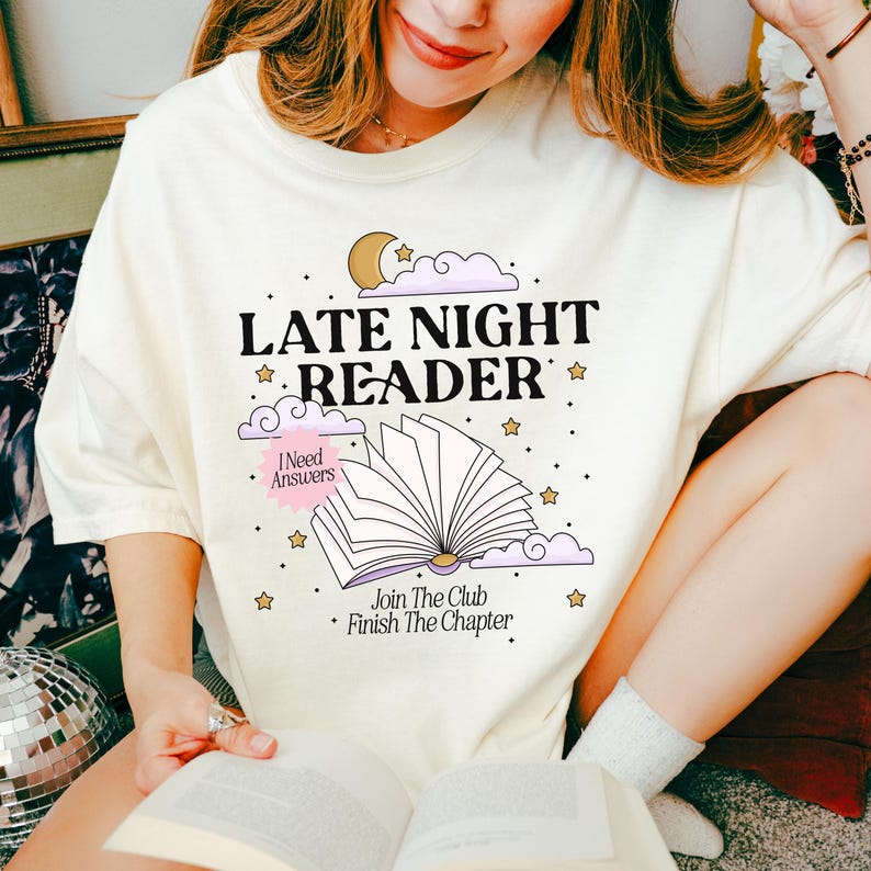 May include: Cream-colored t-shirt with the text "LATE NIGHT READER" in black, with a graphic of an open book, clouds, stars, and a moon. The shirt also has the phrases "I Need Answers" and "Join The Club Finish The Chapter".