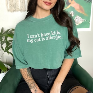 May include: A green t-shirt with the text "I can't have kids, my cat is allergic" in white. The shirt is a solid color and has a relaxed fit. The person is wearing black shorts.
