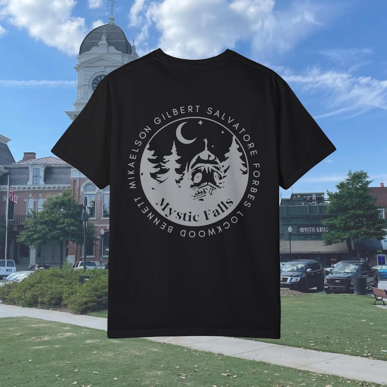 Puede incluir: Camiseta negra con un gr&aacute;fico circular blanco en la espalda. El gr&aacute;fico presenta una luna creciente, estrellas, &aacute;rboles y una casa con el texto "Mystic Falls" y los nombres de los personajes principales del programa de televisi&oacute;n "The Vampire Diaries".