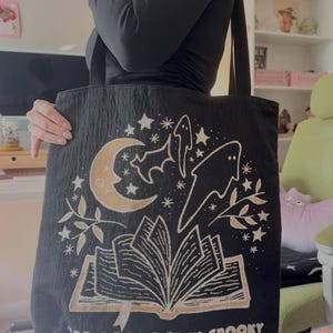 Puede incluir: Bolso tote negro con un dise&ntilde;o espeluznante que presenta una luna creciente, estrellas, murci&eacute;lagos, fantasmas y un libro abierto. El texto "BAD BOOKS STAY SPOOKY" est&aacute; impreso en la parte inferior. El bolso tiene asas negras.