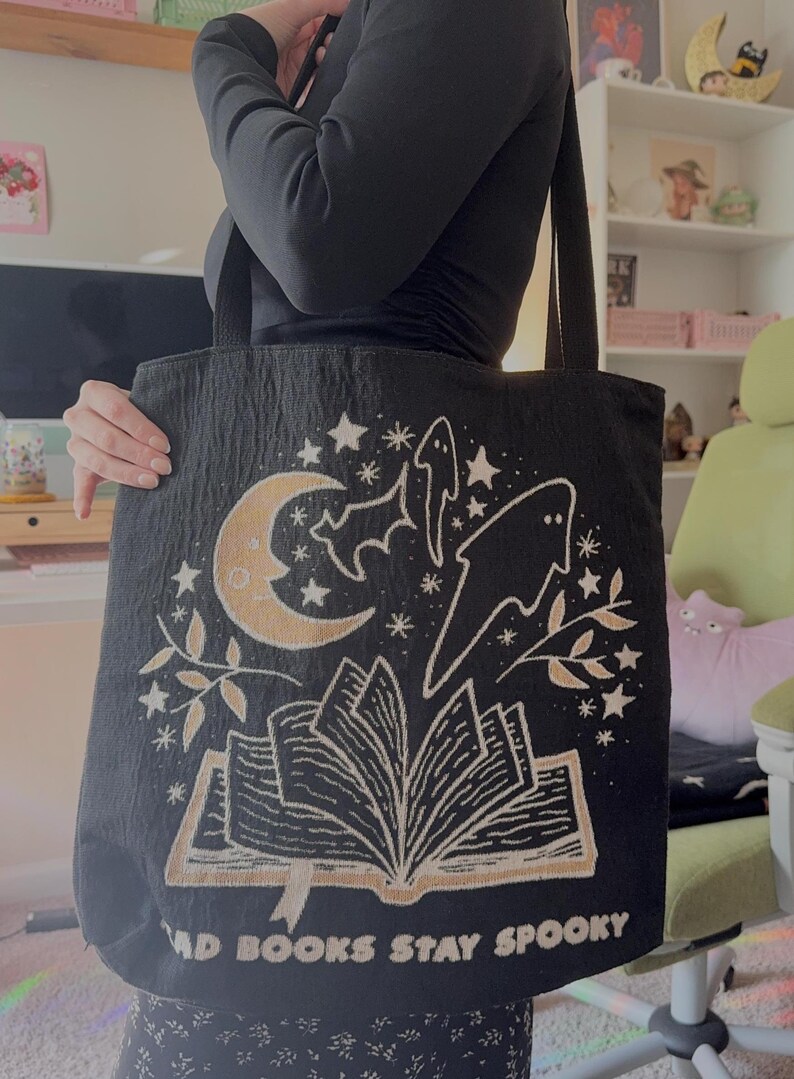 Puede incluir: Bolso tote negro con un dise&ntilde;o espeluznante que presenta una luna creciente, estrellas, murci&eacute;lagos, fantasmas y un libro abierto. El texto "BAD BOOKS STAY SPOOKY" est&aacute; impreso en la parte inferior. El bolso tiene asas negras.