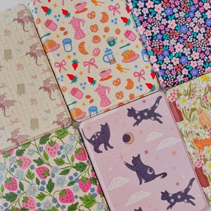 May include: A collection of decorative notebooks with various patterns. Designs include floral, strawberries, dragons, cats with moons and stars, and breakfast-themed illustrations. The notebooks have a rectangular shape and are likely for writing or journaling.