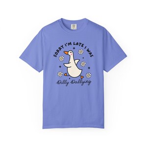 May include: A periwinkle blue t-shirt featuring a cartoon duck with orange feet and beak, surrounded by daisies and stars. The text "SORRY I'M LATE I WAS" and "Dilly Dallying" are in black script.