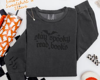 Puede incluir: Una sudadera gris oscuro con el texto "stay spooky read books" en fuente gótica. La sudadera tiene un gráfico de murciélago y siluetas de murciélagos en la manga. Decoraciones de Halloween rodean la sudadera.