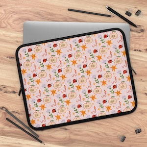 May include: A rectangular laptop sleeve with a light pink background featuring a repeating pattern of red ladybugs, orange star shapes with faces, sliced apples, light purple flowers, and pink worms, finished with a black zipper border.