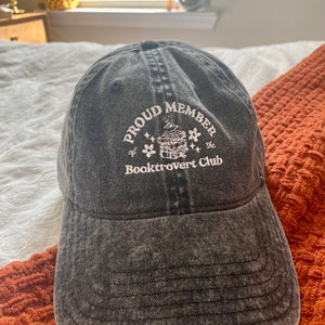 May include: A black denim baseball cap with white embroidery that reads "Proud Member of the Booktrovert Club". The embroidery features a small illustration of a stack of books with stars.