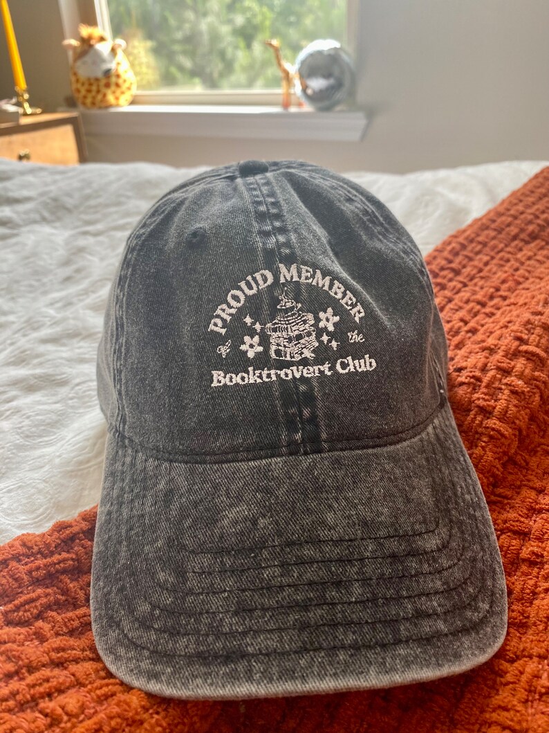 May include: A black denim baseball cap with white embroidery that reads "Proud Member of the Booktrovert Club". The embroidery features a small illustration of a stack of books with stars.