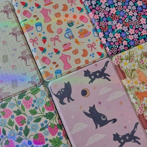 May include: A collection of patterned cases, featuring designs like floral, strawberries, cats with stars, and breakfast items. The cases have a glossy finish and a variety of colors, including pink, blue, and beige.