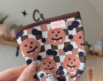 May include: A fabric drink sleeve with a repeating pattern of smiling jack-o'-lantern faces and dark blue cloud shapes on a brown and peach chequered background. The top edge is trimmed with brown fabric.