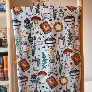 Puede incluir: Una manta suave y azul con un patrón caprichoso de botellas de pociones, calderos, hongos, estrellas y lunas. El patrón se repite en toda la manta.