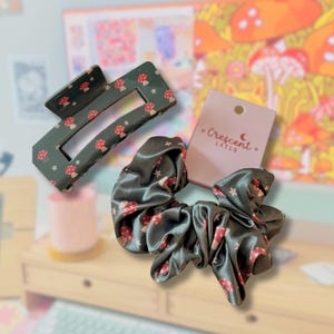 May include: A dark green rectangular hair claw clip and a matching dark green satin scrunchie, both featuring a repeating pattern of small red mushrooms and white stars. A light pink rectangular display card with the text Crescent + Catco.