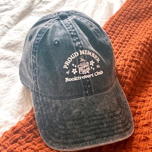 May include: A black denim baseball cap with white embroidery that reads "Proud Member of the Booktrovert Club". The embroidery also includes a stack of books with a star on top.