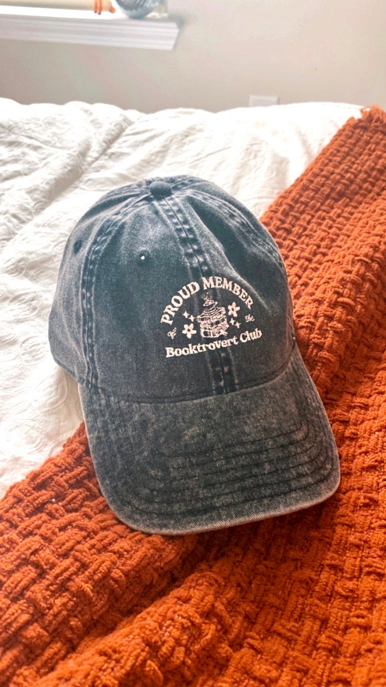 May include: A black denim baseball cap with white embroidery that reads "Proud Member of the Booktrovert Club". The embroidery also includes a stack of books with a star on top.