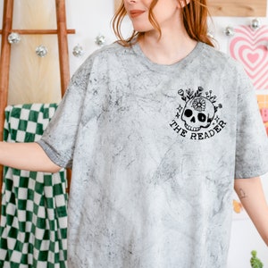 May include: A gray t-shirt with a distressed, marbled look. The shirt features a black and white graphic of a skull with flowers and the text "The Reader".