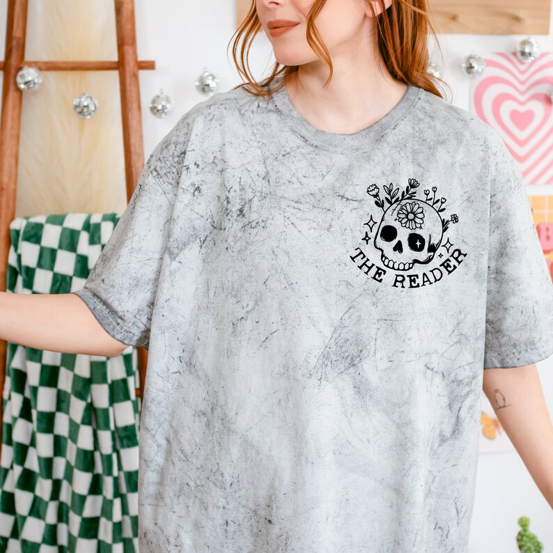 May include: A gray t-shirt with a distressed, marbled look. The shirt features a black and white graphic of a skull with flowers and the text "The Reader".
