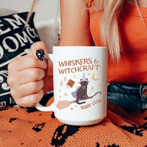 May include: White ceramic mug with the text "WHISKERS & WITCHCRAFT BOOK CLUB". The mug features a black cat wearing a witch hat and cape, riding a broom. Additional illustrations include a book, candles, stars, and a crescent moon.