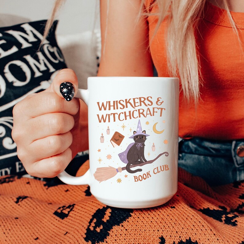May include: White ceramic mug with the text "WHISKERS & WITCHCRAFT BOOK CLUB". The mug features a black cat wearing a witch hat and cape, riding a broom. Additional illustrations include a book, candles, stars, and a crescent moon.