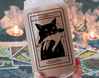 Puede incluir: Una lata de vidrio con una ilustración en blanco y negro de un gato negro sosteniendo un hueso en su boca. La ilustración está rodeada por un borde blanco y el texto "XIII - DEATH" está impreso debajo de la imagen.
