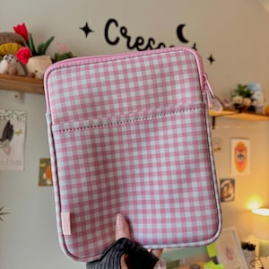 May include: Pink and white gingham pattern tablet sleeve with a pink zipper and a small pink tag. The sleeve has a pocket on the front and is held by a person wearing a black fingerless glove. The background includes a shelf with decor and the word "Crea" in cursive.