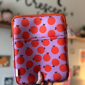 May include: A rectangular, lavender-colored tablet case with a repeating pattern of large, red oranges. The case has an orange zipper and trim, and a small front pocket. The word "Crescent" is written on the wall in cursive.