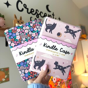May include: Two Kindle Paperwhite II Gen (6.8in) cases. One case has a floral pattern with pink, blue, and white flowers. The other case is light pink with black cat illustrations, stars, and a crescent moon.