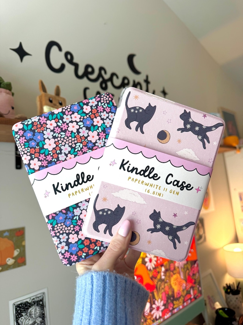 May include: Two Kindle Paperwhite II Gen (6.8in) cases. One case has a floral pattern with pink, blue, and white flowers. The other case is light pink with black cat illustrations, stars, and a crescent moon.