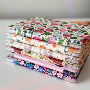 May include: A stack of floral patterned phone cases. The cases feature various designs, including strawberries, flowers, and other botanical elements. The cases have clear edges and a variety of colors, including pink, blue, and green.
