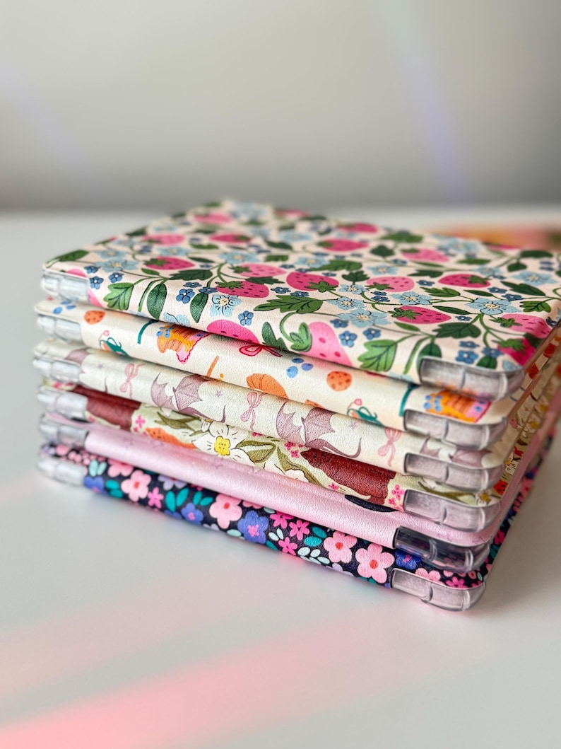 May include: A stack of floral patterned phone cases. The cases feature various designs, including strawberries, flowers, and other botanical elements. The cases have clear edges and a variety of colors, including pink, blue, and green.