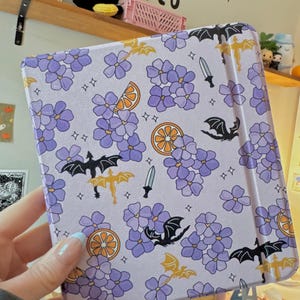 May include: A rectangular tablet case with a light purple background featuring a repeating pattern of purple flower clusters, orange citrus slices, small black and gold dragon silhouettes, silver swords, and tiny four-pointed star accents.