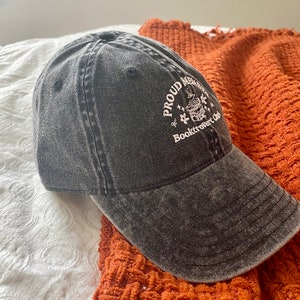 May include: A black denim baseball cap with white embroidery that reads "Proud Member of Booktrovert Club" and features a small illustration of a book with a stack of books on top.