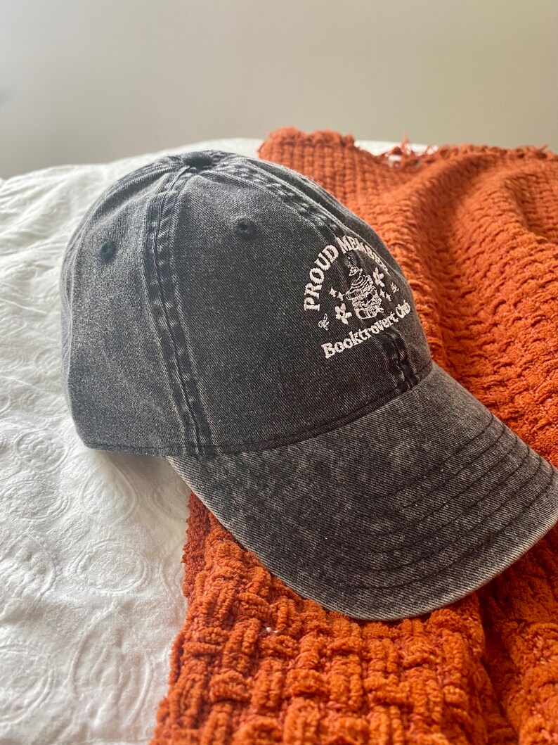 May include: A black denim baseball cap with white embroidery that reads "Proud Member of Booktrovert Club" and features a small illustration of a book with a stack of books on top.