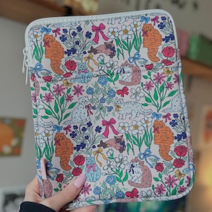 May include: A light-colored laptop sleeve with a colorful floral and cat pattern. The design features orange, blue, and pink cats among flowers and bows. The sleeve has a white zipper and trim.
