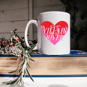 Peut inclure: Mug en céramique blanche avec un motif de cœur rouge et rose. Les mots "Villain Lover" sont imprimés en police script dans le cœur. La tasse a une anse incurvée et est posée sur une pile de livres.