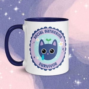 Puede incluir: Taza de cerámica blanca con borde azul. La taza presenta un gato azul con un brote verde en la cabeza y una sola lágrima. El gato está dentro de un círculo con el texto "Social Gathering Survivor" en azul y blanco.