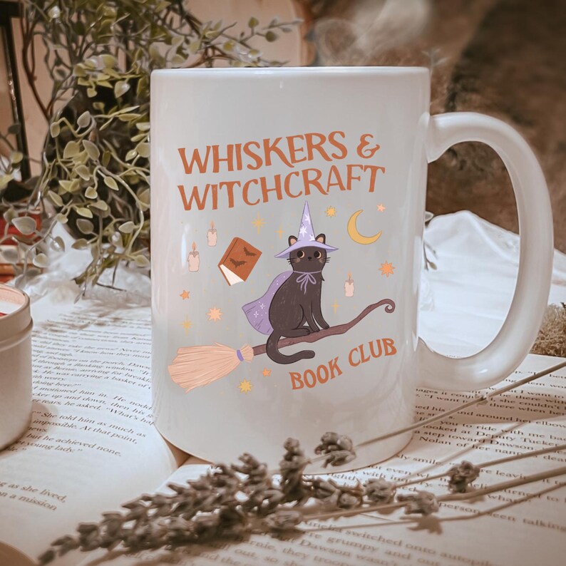 May include: White ceramic mug with the text "WHISKERS & WITCHCRAFT BOOK CLUB". The mug features a cartoon black cat wearing a witch hat and cape, riding a broom. The design includes a book, candles, and stars.