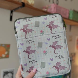 May include: A light gray tablet case with a repeating pattern of purple dragons, butterflies, books, and flowers. The text "Fantasy Reader" is printed on the case. The case has a green zipper and a pink tag.