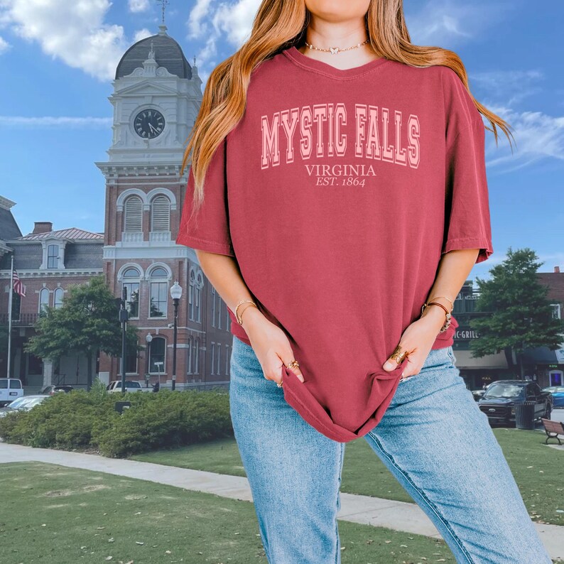 Puede incluir: Una camiseta granate con el texto "Mystic Falls, Virginia Est. 1864" impreso en rosa.